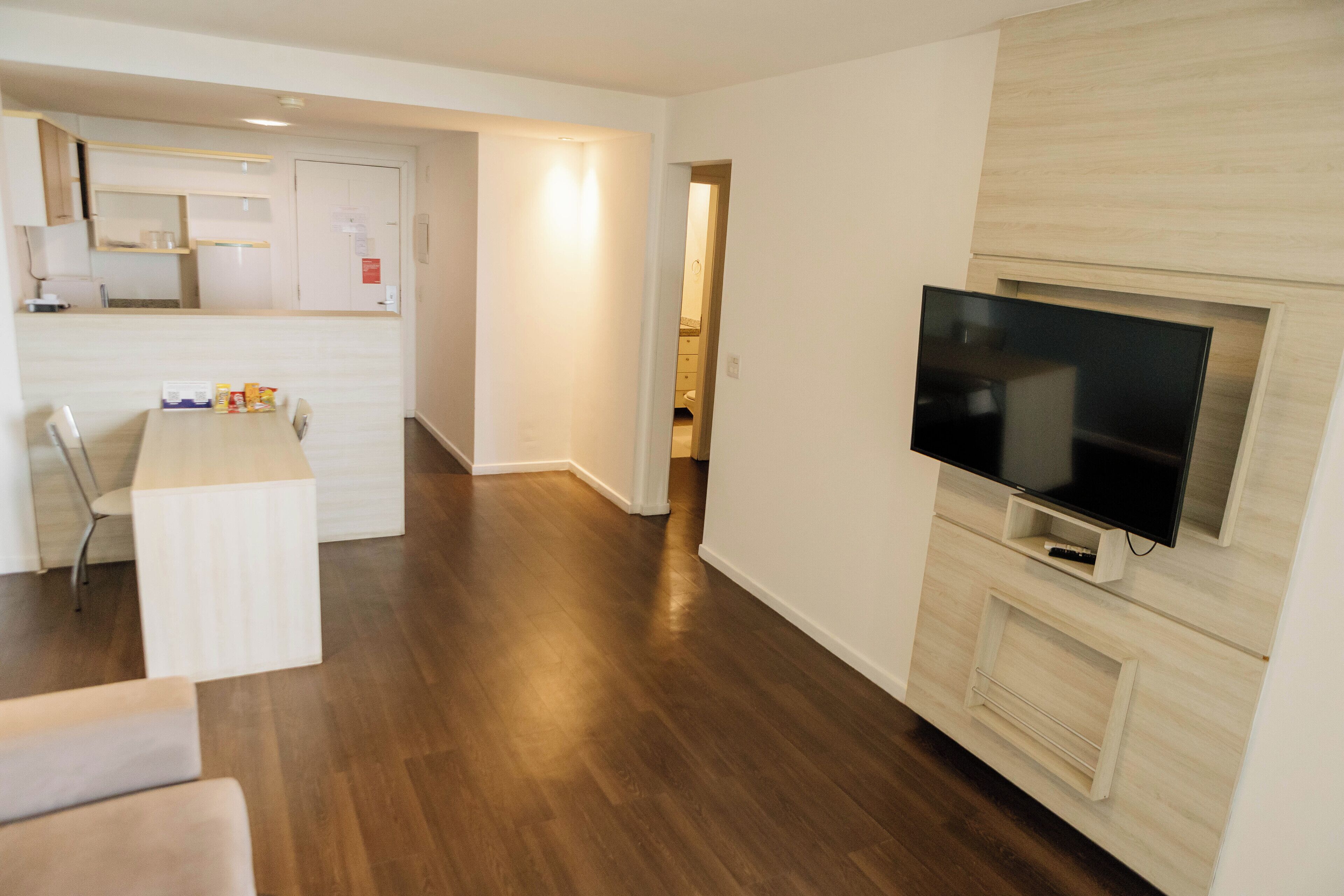 Een moderne woonkamer met een flatscreen televisie aan de muur, een eethoek met tafel en stoelen, en een keuken met kasten en een aanrecht.