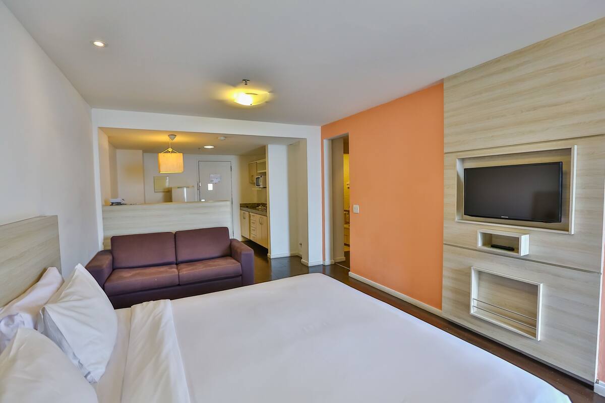 A hotel room with a large bed, a sofa, a TV mounted on the wall, and a kitchenette in the background.