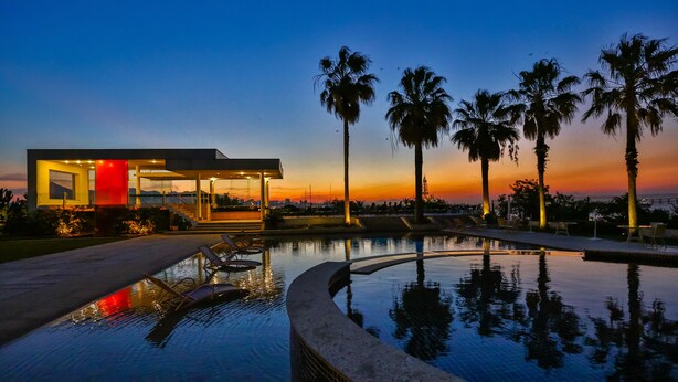 A modern building with a pool, palm trees, and a sunset view.
