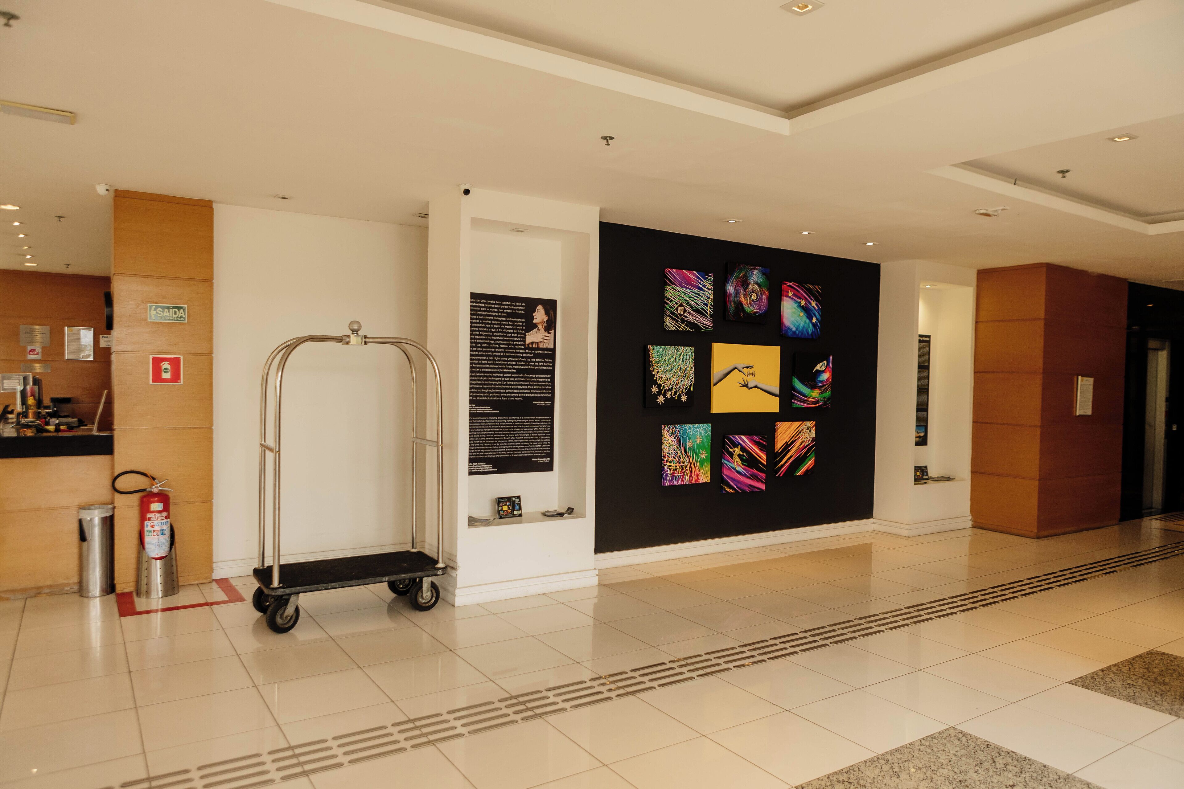 Un vestíbulo de hotel con un diseño moderno y limpio, que presenta un muro con ilustraciones coloridas, un extintor, un carrito y un cartel que indica la ubicación.