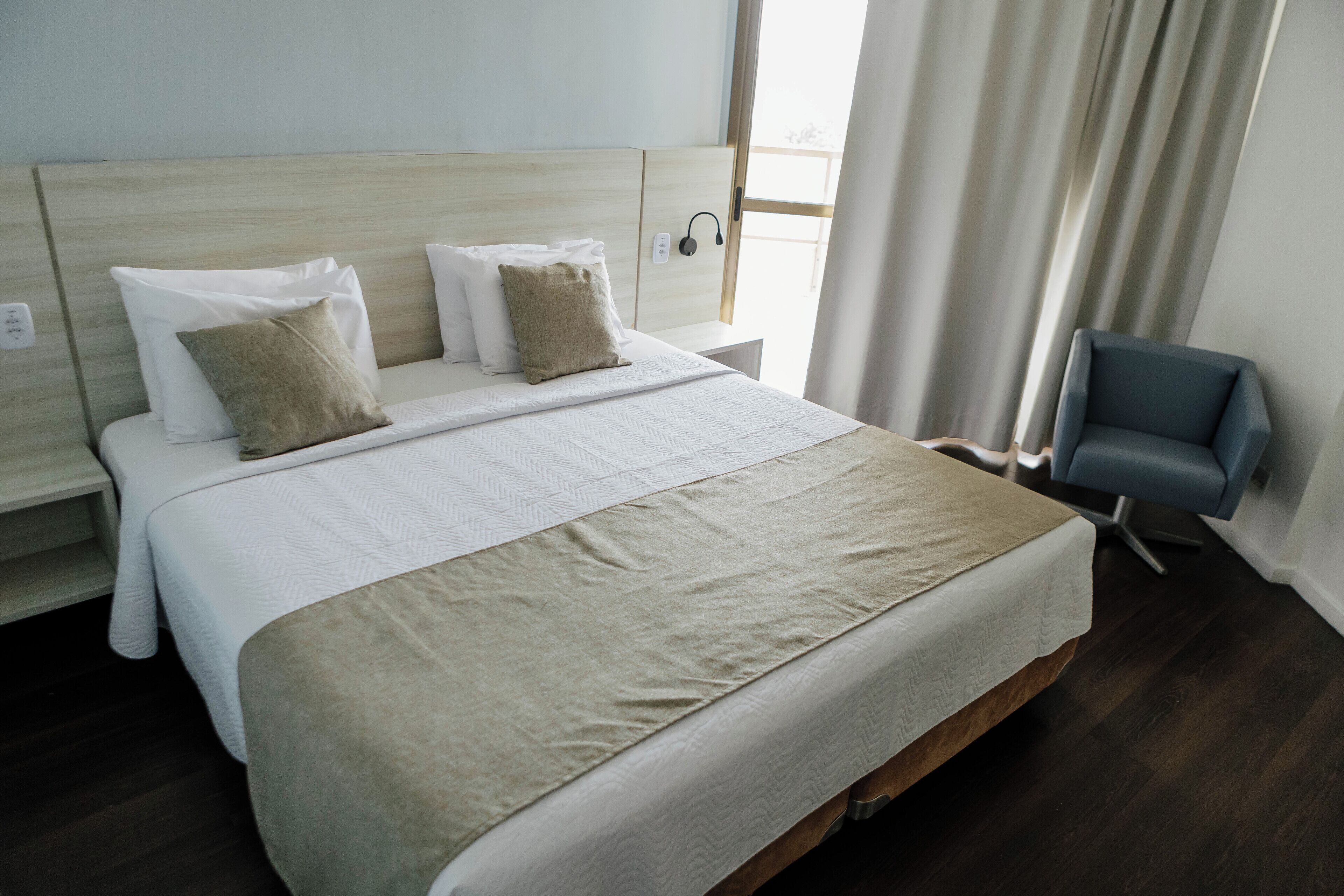 Una cama bien hecha con una colcha color beige y almohadas, un cabecero de madera, una mesita de noche con una lámpara y una silla.