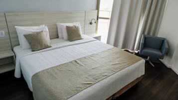 Una cama bien hecha con una colcha color beige y almohadas, un cabecero de madera, una mesita de noche con una lámpara y una silla.