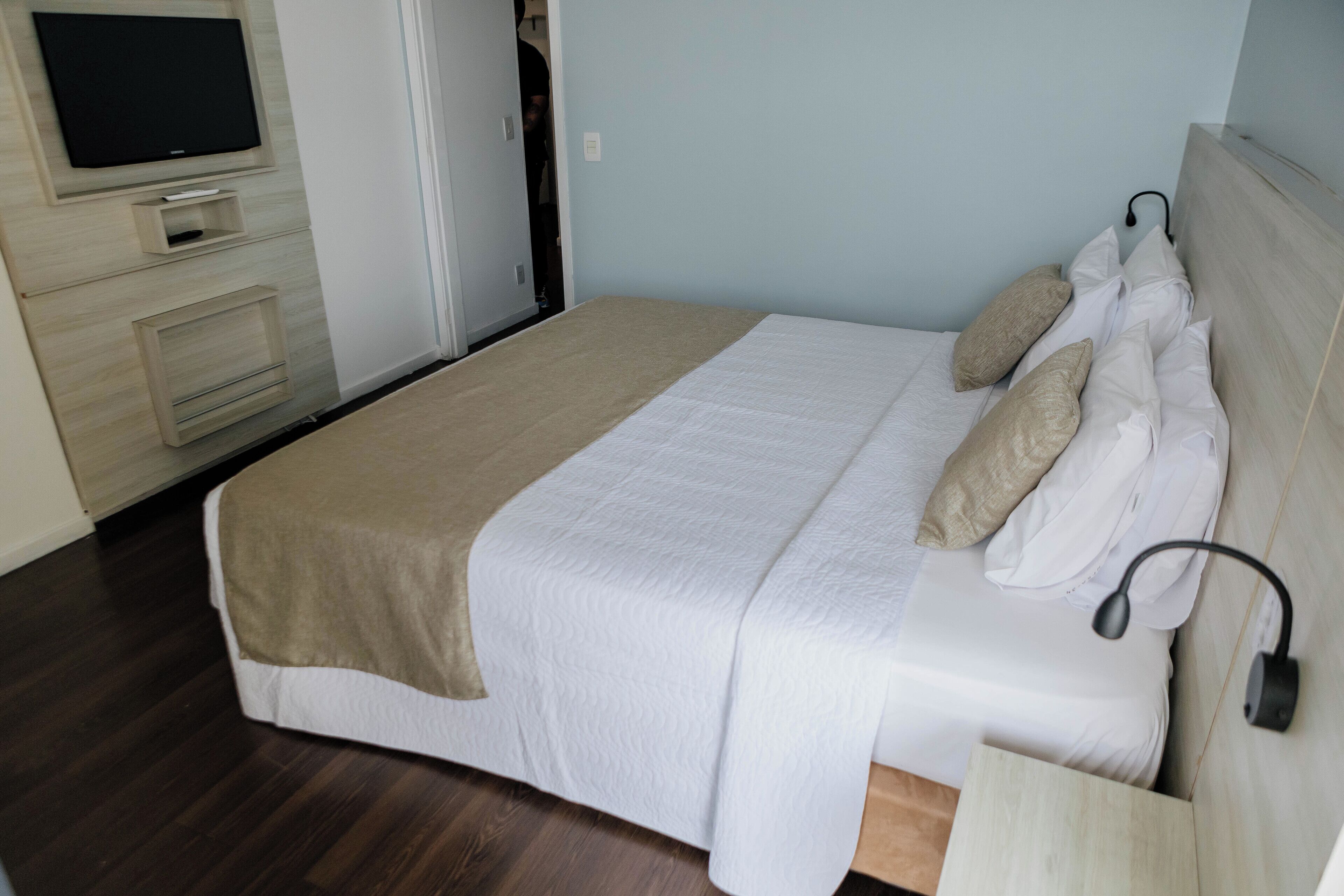 A neatly made bed with a white and beige bedspread, a bedside lamp, and a flat-screen TV mounted on the wall above the bed.