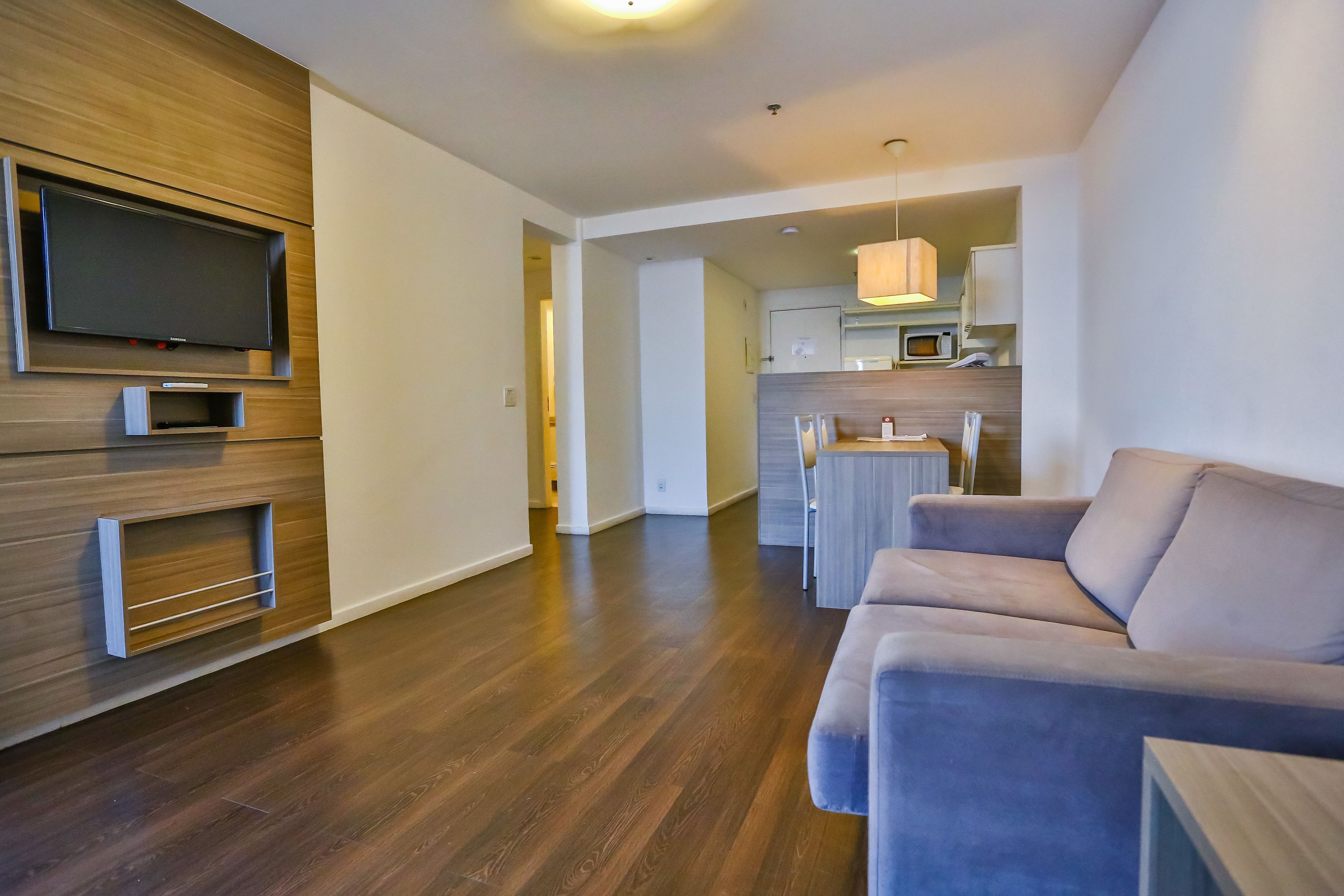A modern living room with a flat-screen TV, wooden paneling, a grey sofa, and a kitchen area in the background.