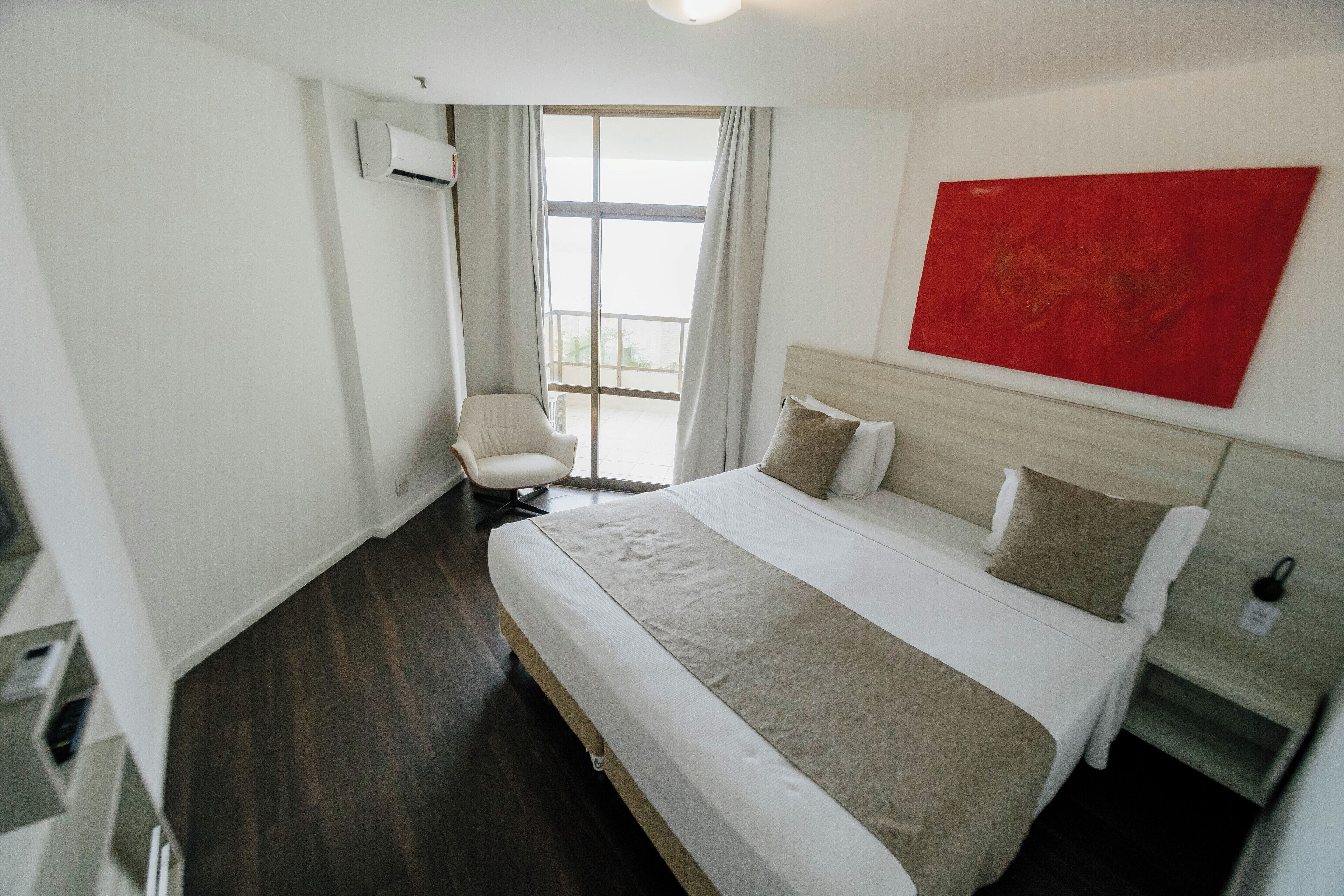 A hotel room with a large bed, a chair, a red artwork on the wall, and a window with curtains.