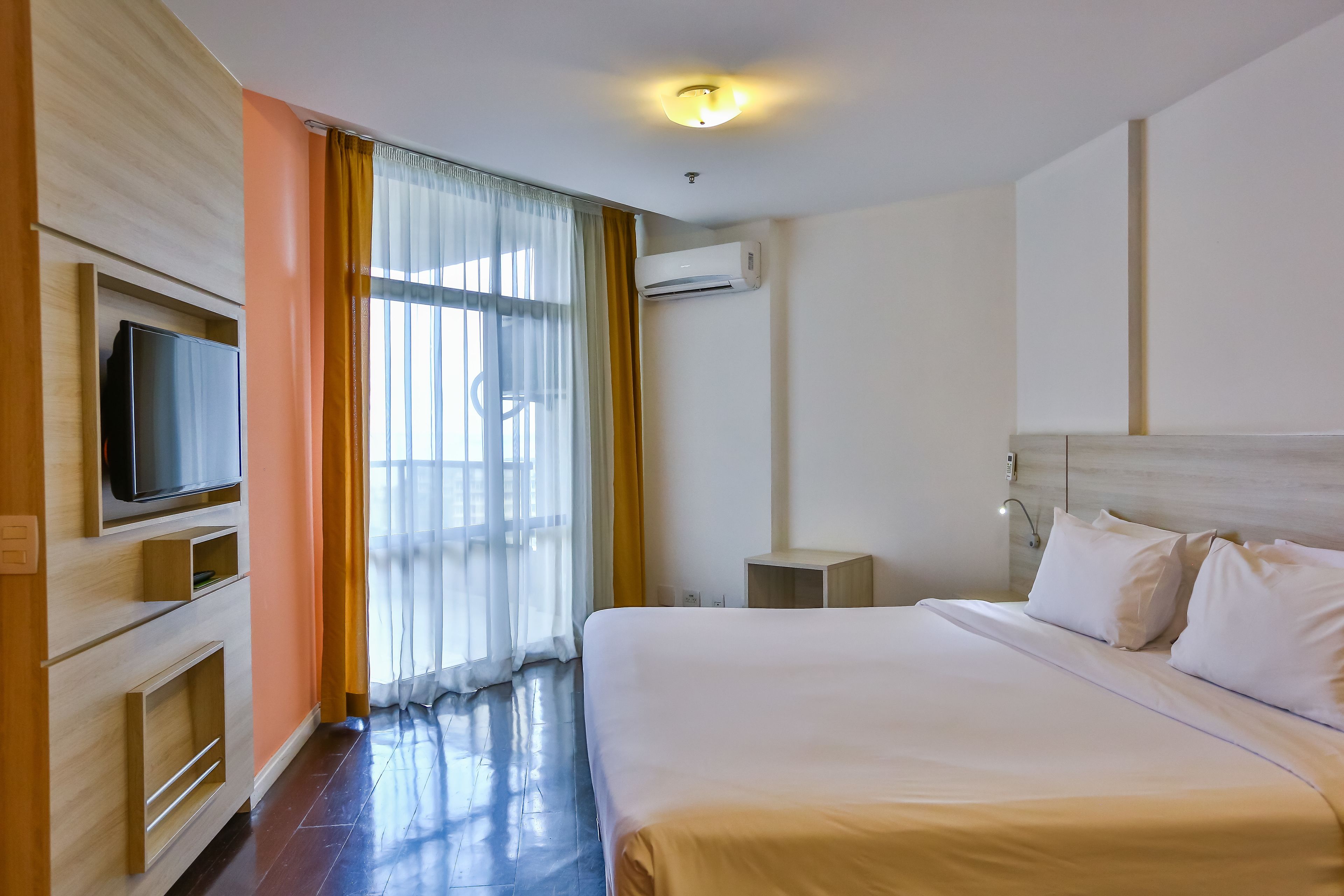 A hotel room with a bed, a TV, an air conditioning unit, and a window with curtains.