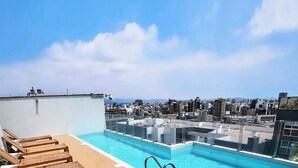 Une piscine sur le toit offrant une vue sur la ville, des chaises longues en bois et une barrière en verre.