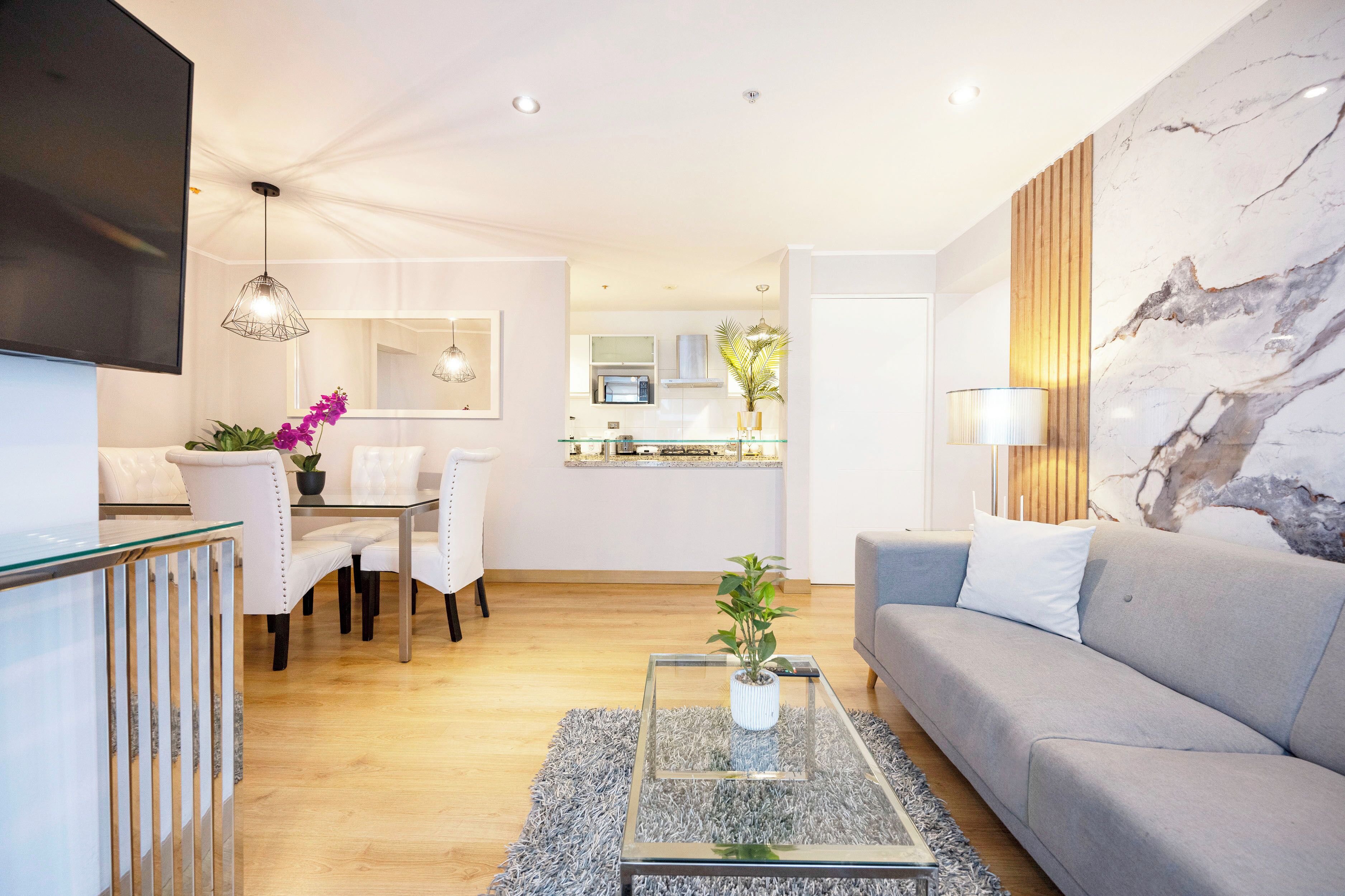 Una sala de estar moderna con un sofá gris, una mesa de centro de vidrio y un comedor con sillas blancas.