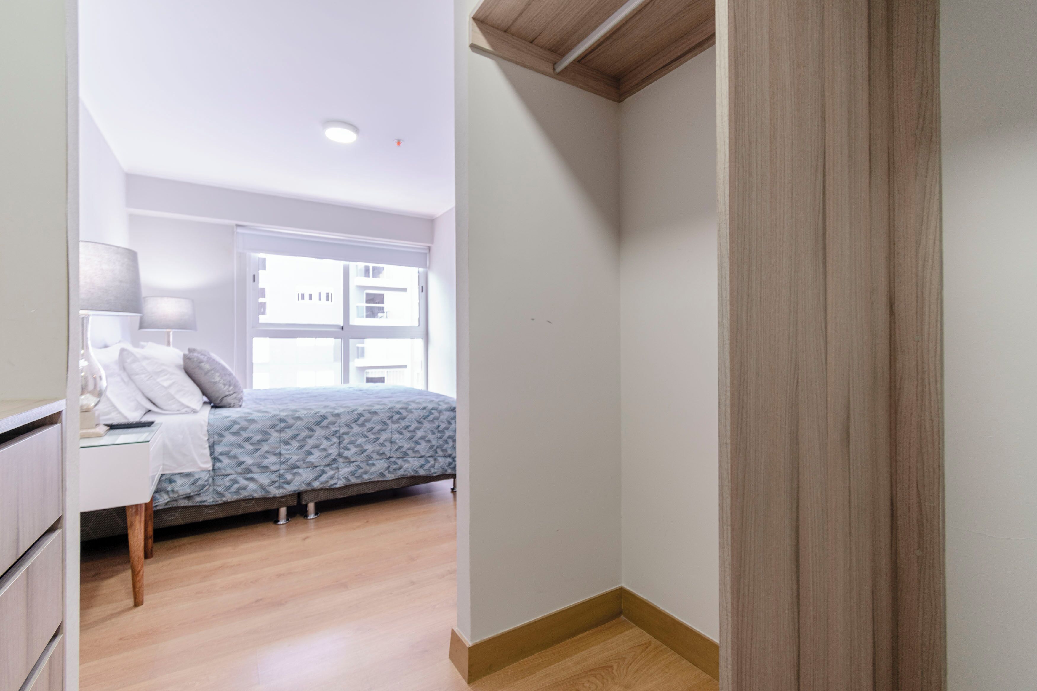 Un dormitorio con cama, mesita de noche, lámpara y ventana con vistas a edificios.