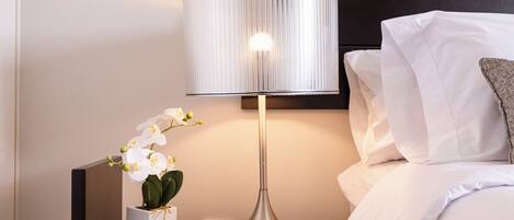 Ein ordentlich bezogenes Bett mit weißen Leinen, ein Nachttisch mit Lampe und eine Orchidee im Topf.