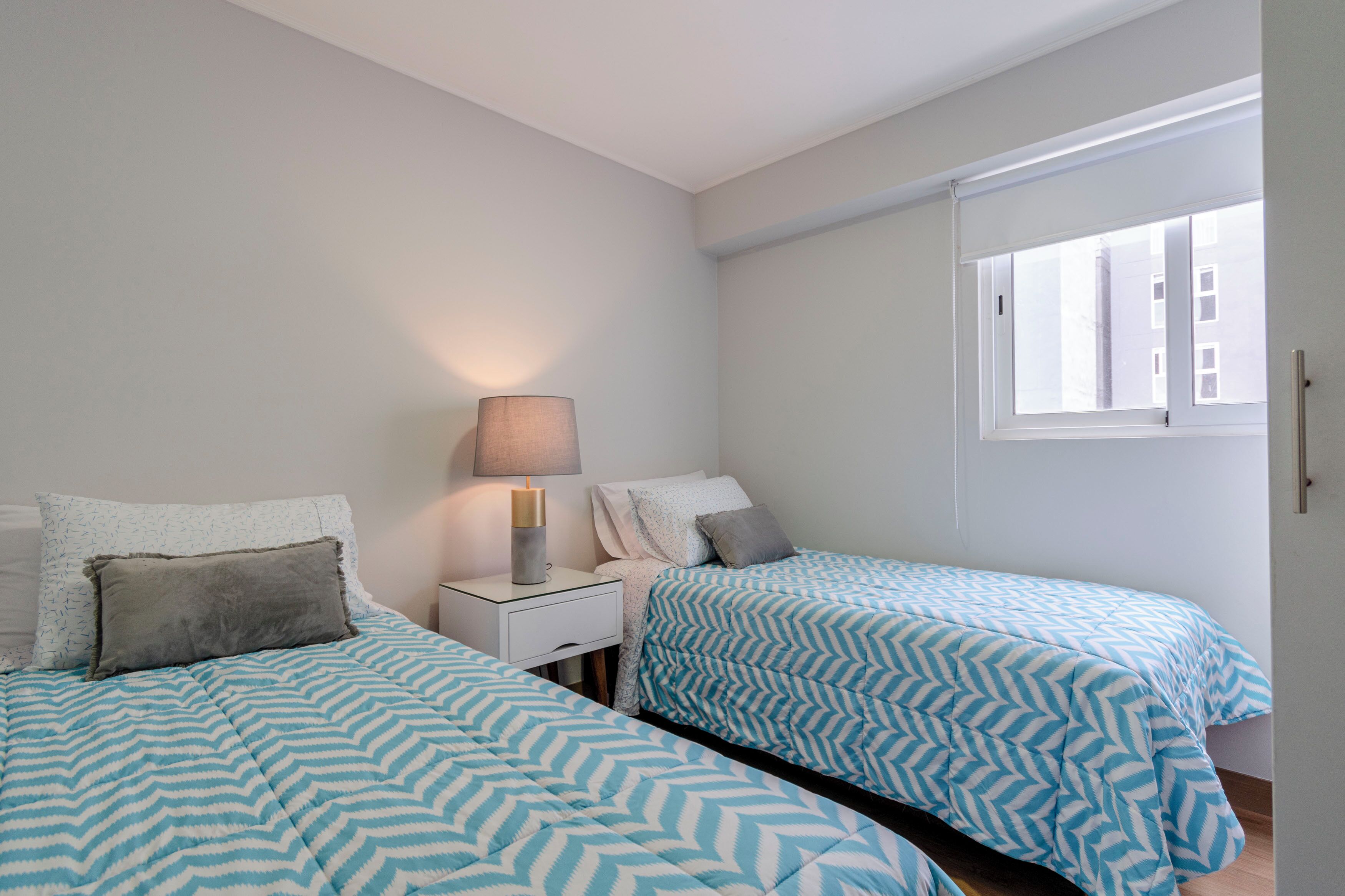 Un dormitorio con dos camas, una mesita de noche con lámpara y una ventana con persianas.
