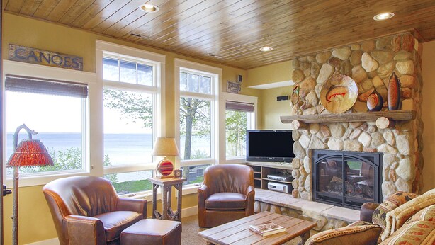 A cozy living room with leather chairs, a stone fireplace, a television, and a view of the ocean through large windows. - Trappers Landing Lodge (Walker)