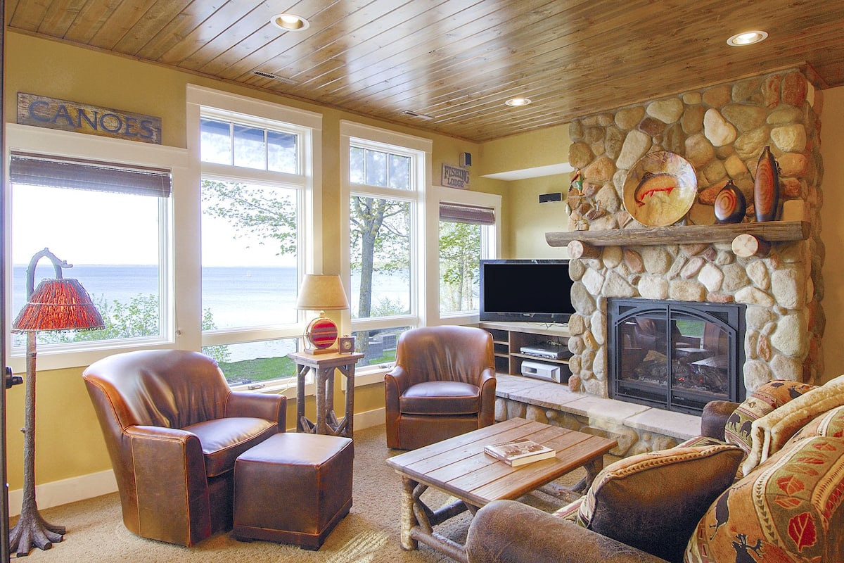 A cozy living room with leather chairs, a stone fireplace, a television, and a view of the ocean through large windows.