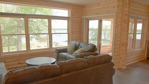A cozy living room with a brown leather sofa, a round coffee table, and large windows offering a view of a lake and trees. - Trappers Landing Lodge (Walker)