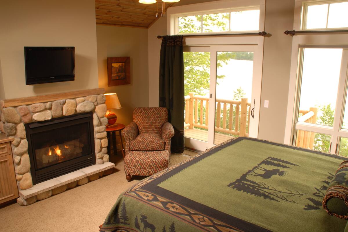 A bedroom with a stone fireplace, a patterned armchair, a bed with a green blanket, and a balcony with a wooden railing.