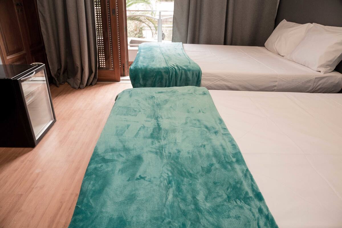Une chambre d’hôtel avec un lit, un banc, un plancher en bois et une fenêtre avec des rideaux.