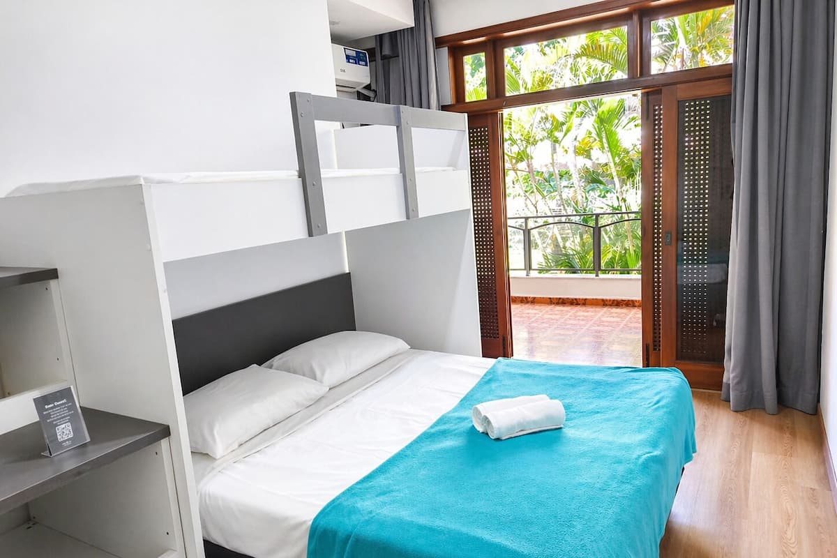 Un lit simple avec un matelas blanc et une couverture turquoise, une petite table de chevet et une vue sur la verdure à travers une porte vitrée.