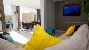 Una sala moderna con un sofá, cojines amarillos y negros, un televisor montado en la pared y un comedor con sillas y una mesa.