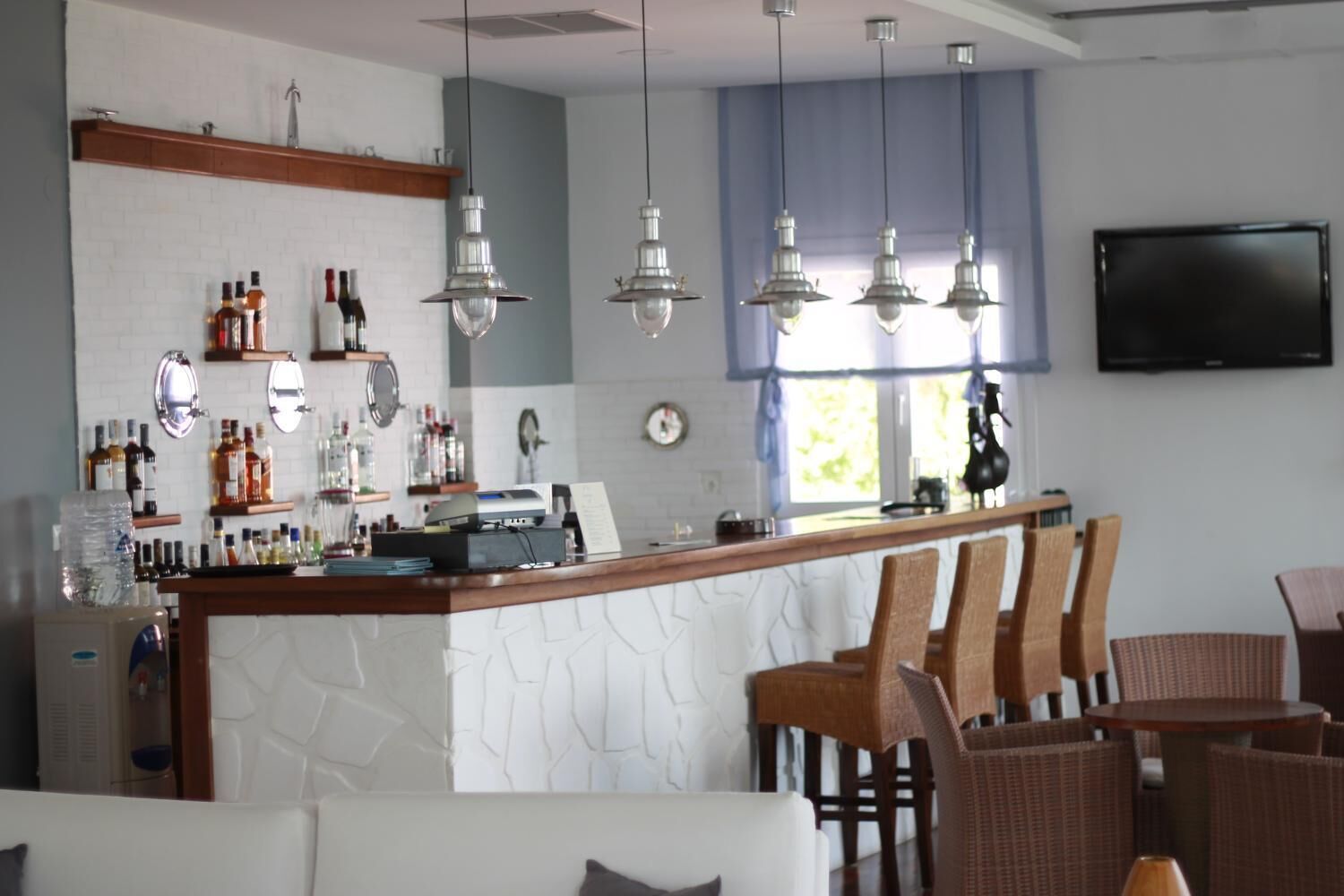 Uma área de bar com balcão, banquetas e luminárias pendentes. Estantes exibem garrafas e há uma televisão fixada na parede.