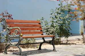 Un banc en bois doté d’accoudoirs et d’un dossier en métal, placé sur une surface pavée, avec un ciel dégagé et quelques feuillages en arrière-plan.
