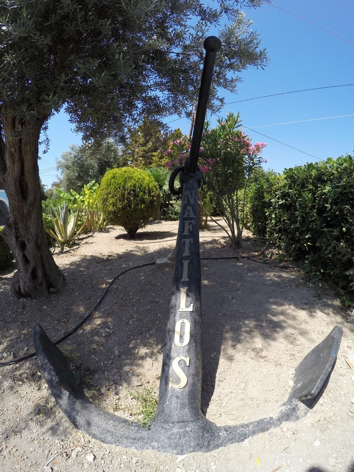 An anchor-shaped sculpture with the inscription "NAFILOS" in a garden setting.