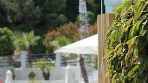 A pool area with a wooden structure for a shower, surrounded by lounge chairs and umbrellas. - Naftilos (Samos)
