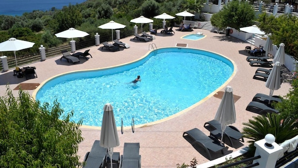 A swimming pool with lounge chairs and umbrellas, surrounded by greenery and overlooking a body of water. - Naftilos (Samos)
