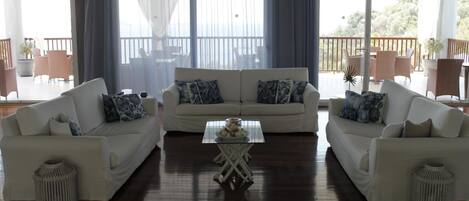 A spacious living room with a central coffee table, two white sofas with patterned cushions, and a large window with sheer curtains.