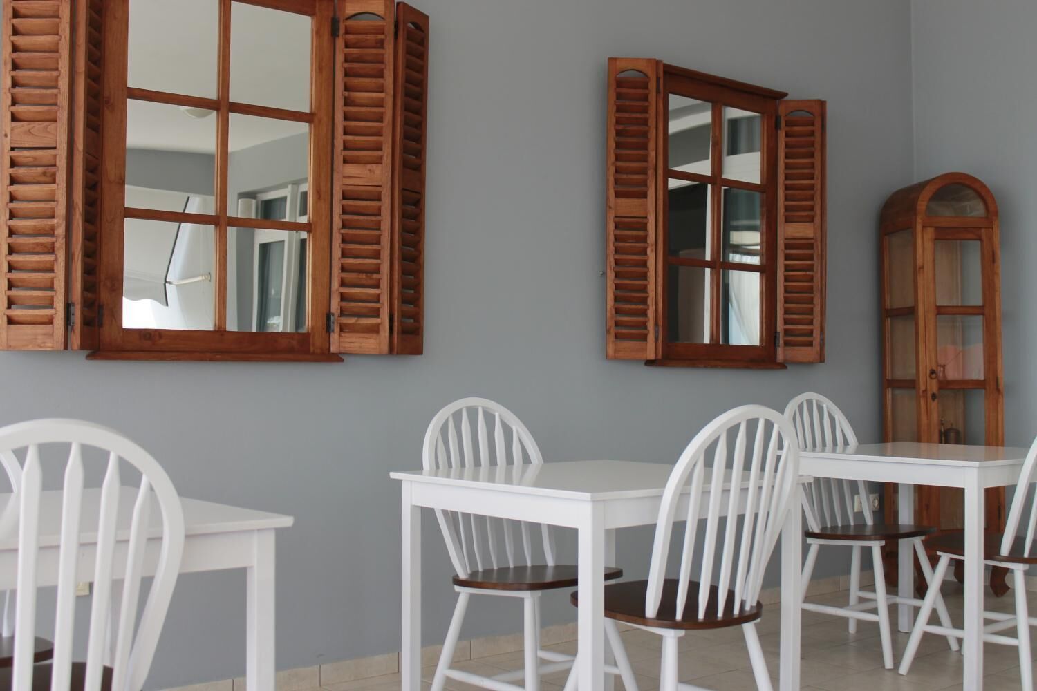 A dining area with white chairs and a table, wooden shutters on the windows, and a tall cabinet.