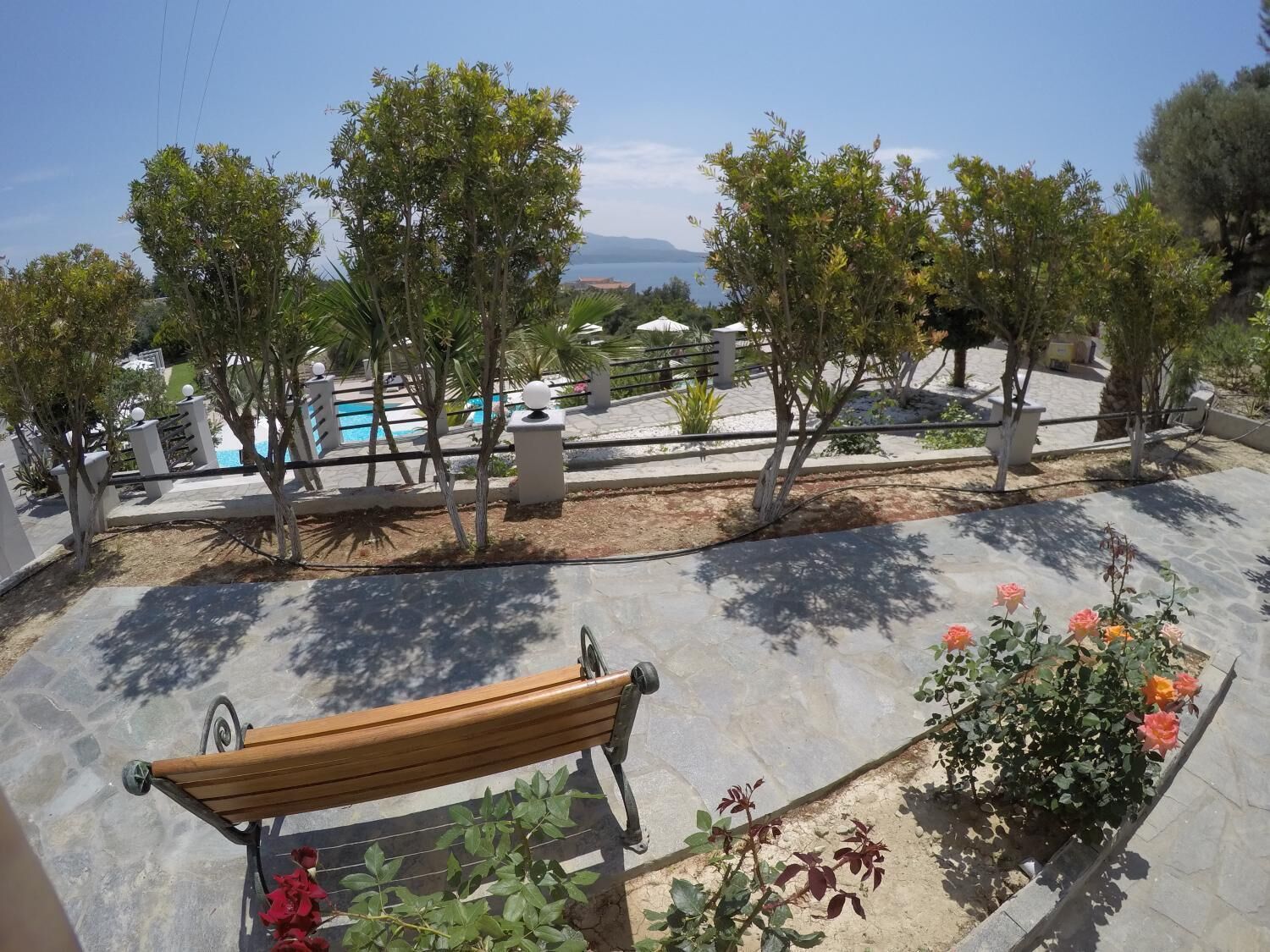 A patio area with a wooden bench, a flower bed with roses, and a view of a pool and mountains in the distance.