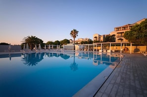A swimming pool with lounge chairs and umbrellas, surrounded by buildings with balconies and palm trees. - The Bay (Zakynthos)