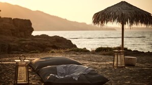 A beach setting with a thatched umbrella, a lantern, and a cushion on the sand.