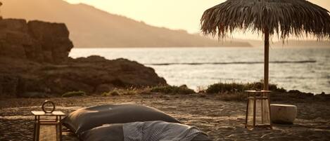A beach setting with a thatched umbrella, a lantern, and a cushion on the sand.
