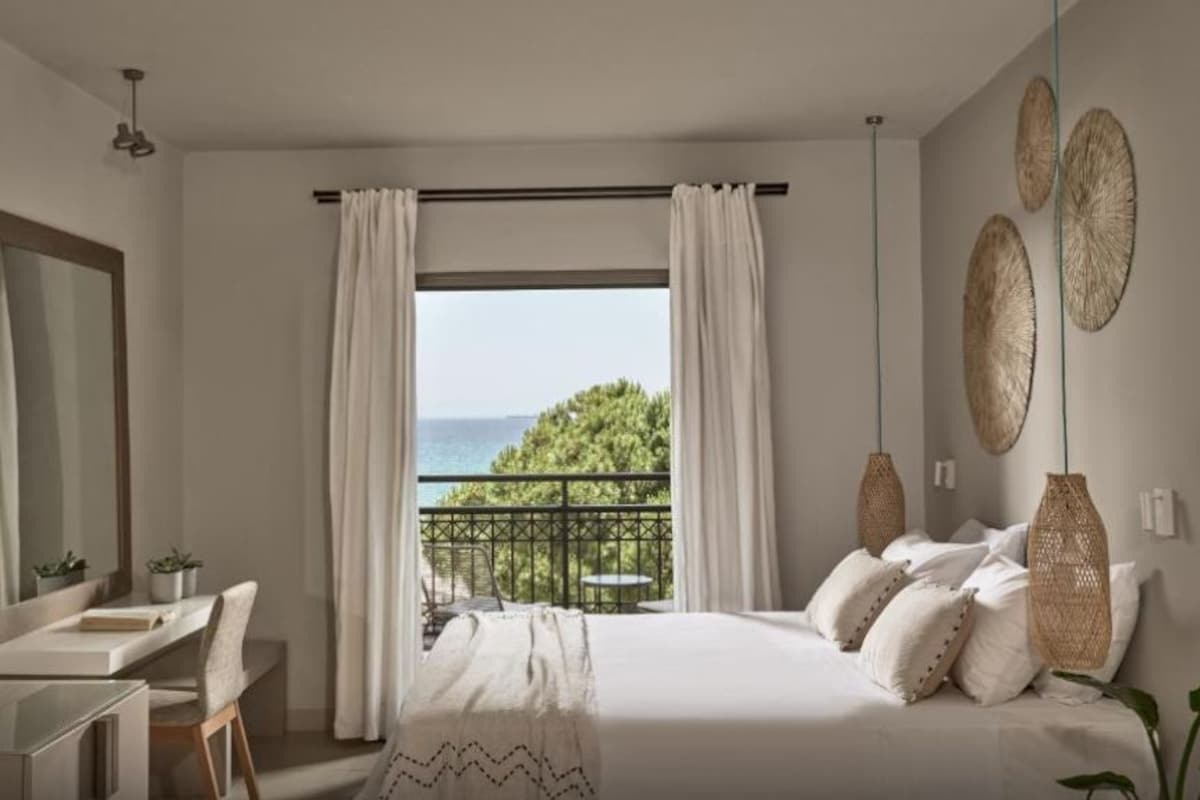 Une chambre avec un lit, un bureau, une chaise, un balcon donnant sur la mer et des œuvres d’art décoratives au mur.