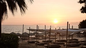 A beach with umbrellas and lounge chairs, a sunset in the background, and a calm sea. - The Bay (Zakynthos)