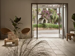 An interior space with a view of an outdoor garden through a glass door, a seating area with chairs, and a table with a plant. - The Bay (Zakynthos)