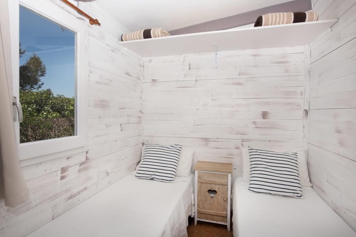 A small, white wooden room with two beds, each with striped pillows, and a small wooden nightstand with a heart-shaped cutout.