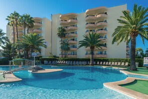 Un hotel con varios balcones, una piscina con sillones de descanso y palmeras.