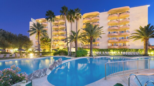 A hotel with a swimming pool, palm trees, and a well-lit building facade. - Aparthotel Alcudia Beach (Alcúdia)
