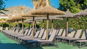 A row of sun loungers under thatched umbrellas, with a green lawn and a hill in the background. - Aparthotel Alcudia Beach (Alcúdia)