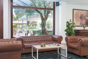A room with a brown leather sofa, a wooden coffee table, and a large window with a view of outdoor greenery and a tree. - Aparthotel Alcudia Beach (Alcúdia)