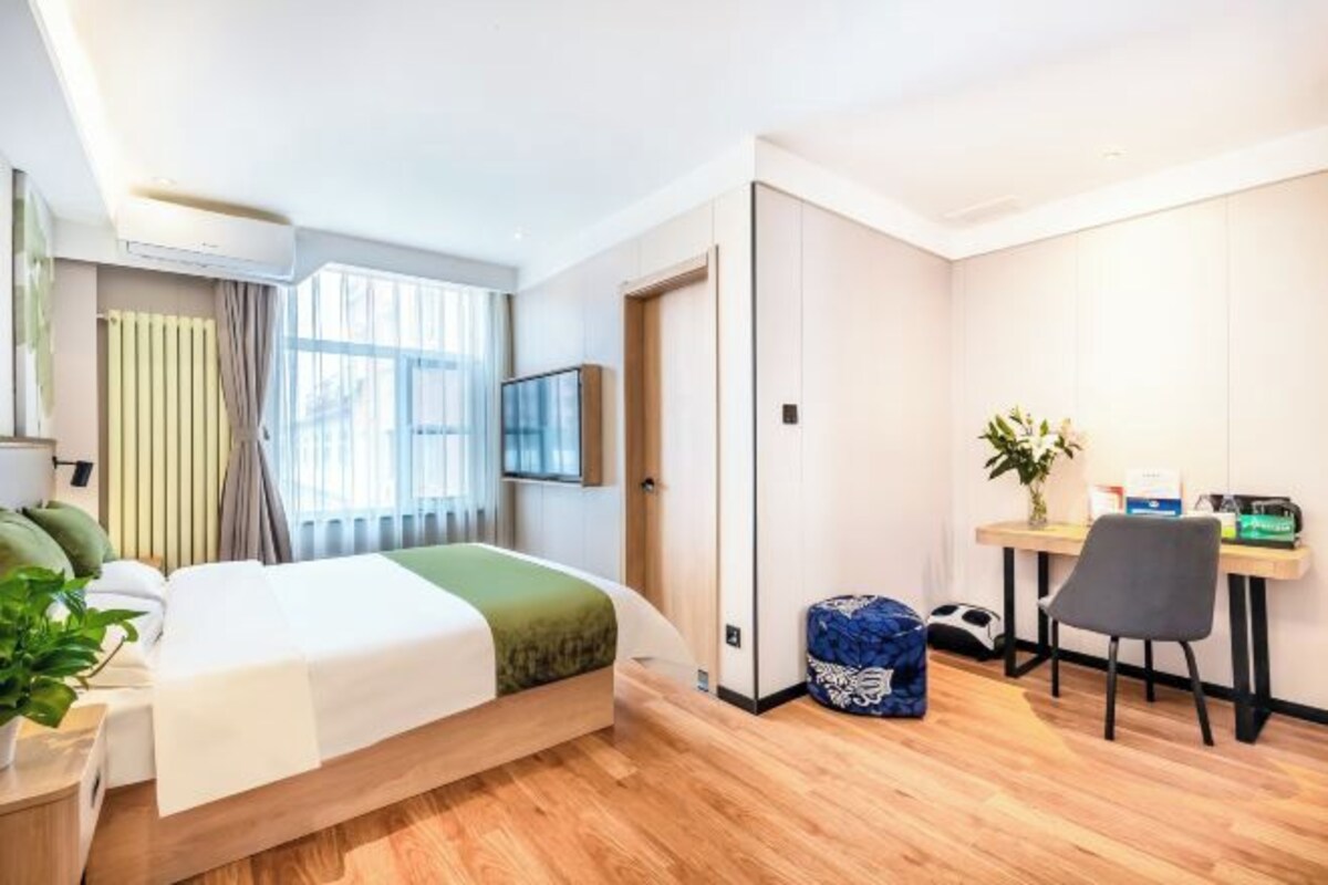 A modern hotel room with a bed, a desk, a chair, a potted plant, and a window with curtains.