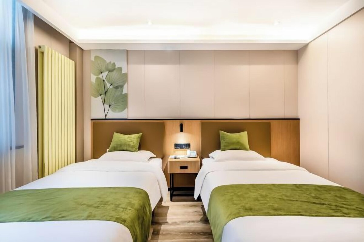 Une chambre d’hôtel avec deux lits, une table de chevet, une lampe fixée au mur et une grande fenêtre avec des rideaux.