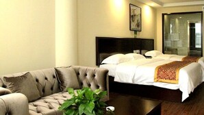 Habitación de hotel con un sofá capitonado, una cama grande con ropa de cama blanca, una mesa de centro de madera oscura y una planta verde.