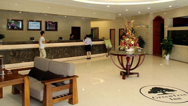 Un vestíbulo de hotel con mostrador de recepción, una zona de estar con banca y una mesa, y un centro de mesa con plantas decorativas.