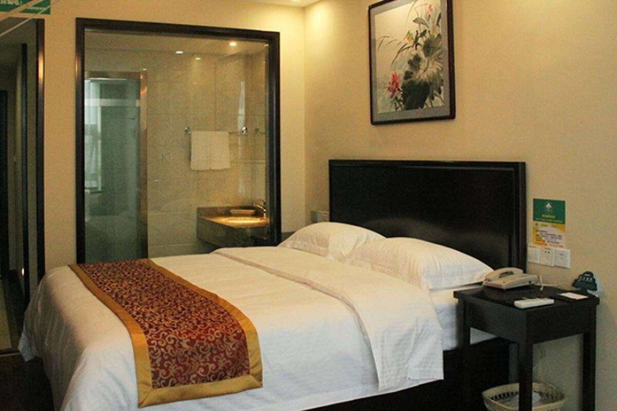 A hotel room with a bed, a bedside table with a phone and a basket, a bathroom with a glass shower enclosure, and a painting on the wall.
