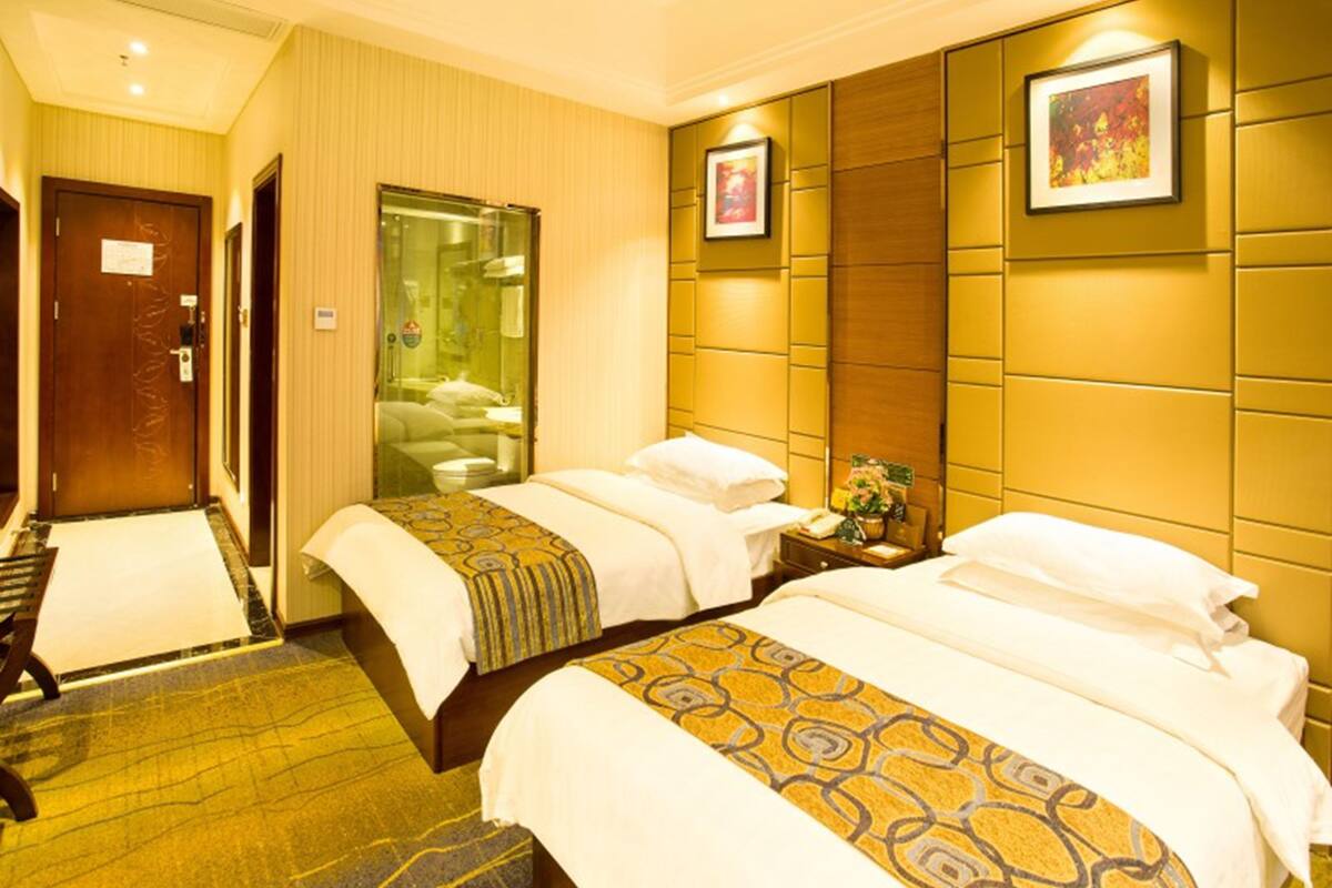 A hotel room with two beds, a bathroom with a glass door, and a wooden headboard.