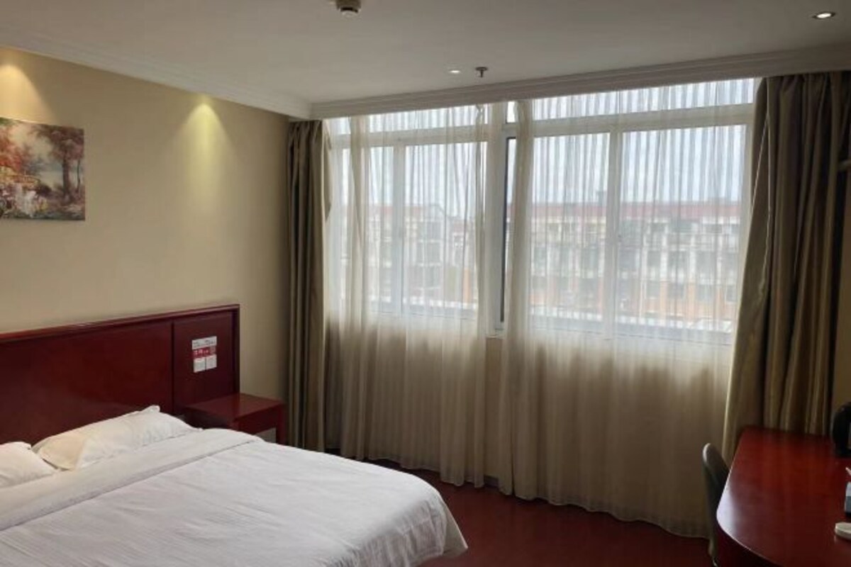 A hotel room with a bed, a nightstand, a desk, and a large window with curtains.