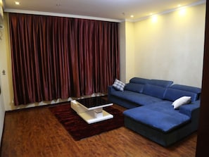 Un salón con un sofá azul, una mesita blanca y una cortina rojo oscuro.
