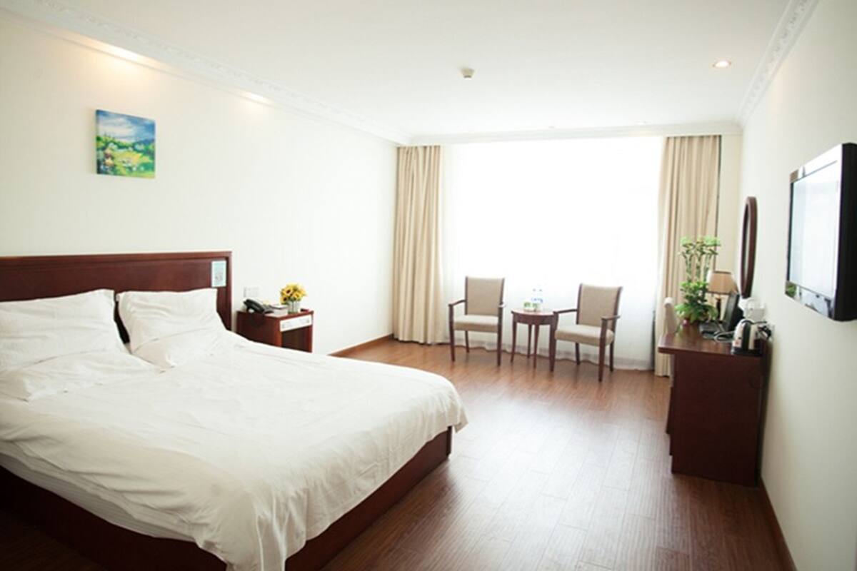 Une chambre d’hôtel avec un grand lit, un bureau avec une chaise, une petite table avec deux chaises, une plante et un téléviseur fixé au mur.